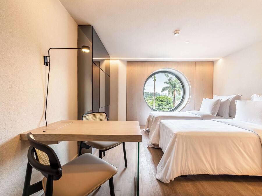 Quarto de hotel moderno com escrivaninha de madeira, duas camas, janela redonda com vista para palmeiras e luminária fixada na parede.