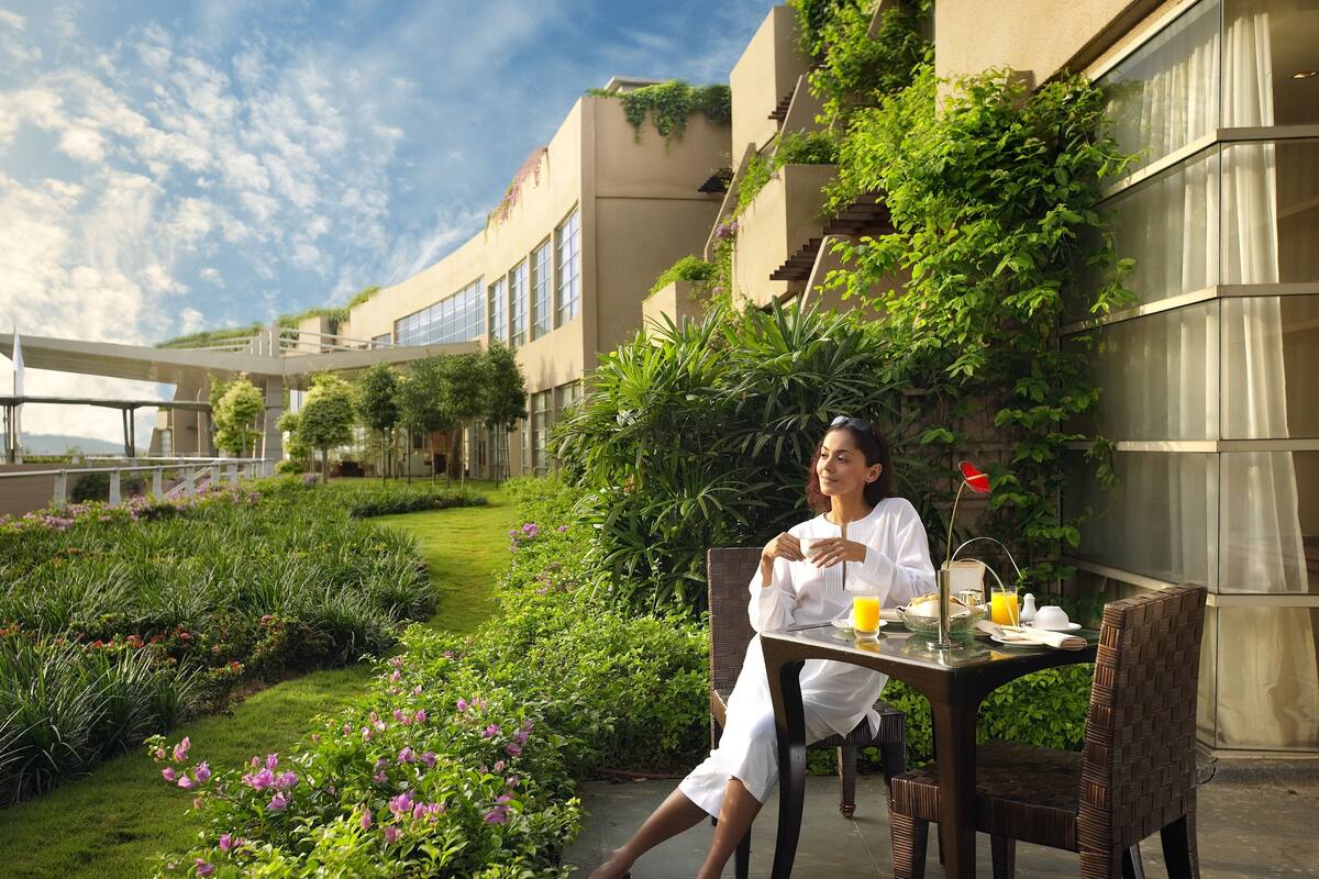 A person sitting at an outdoor table with a drink and a plate of food, surrounded by lush greenery and a building with large windows.