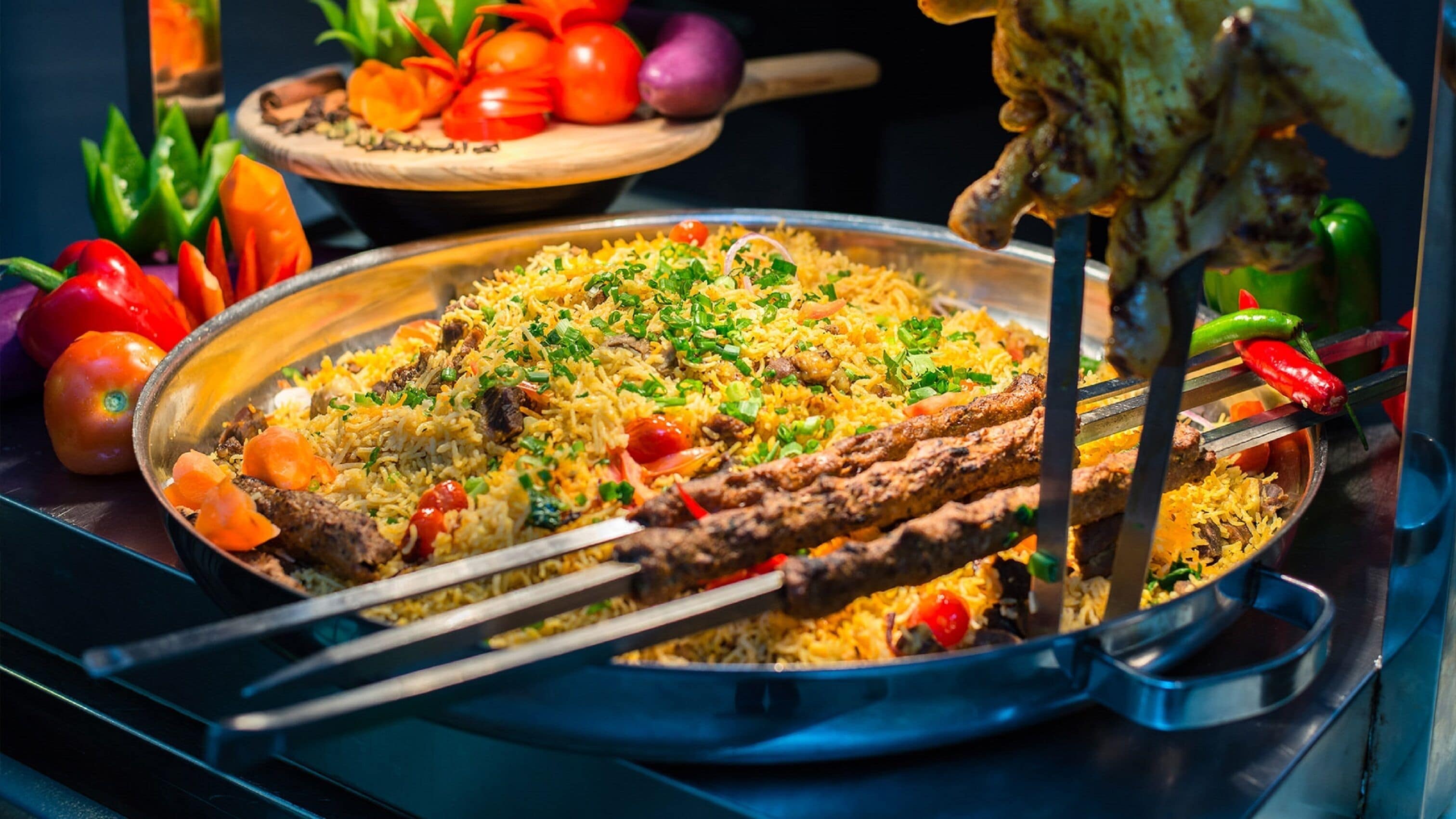 Eine große Schüssel Biryani mit Fleisch und Gemüse, dazu Spieße mit gegrilltem Fleisch und frischem Gemüse.