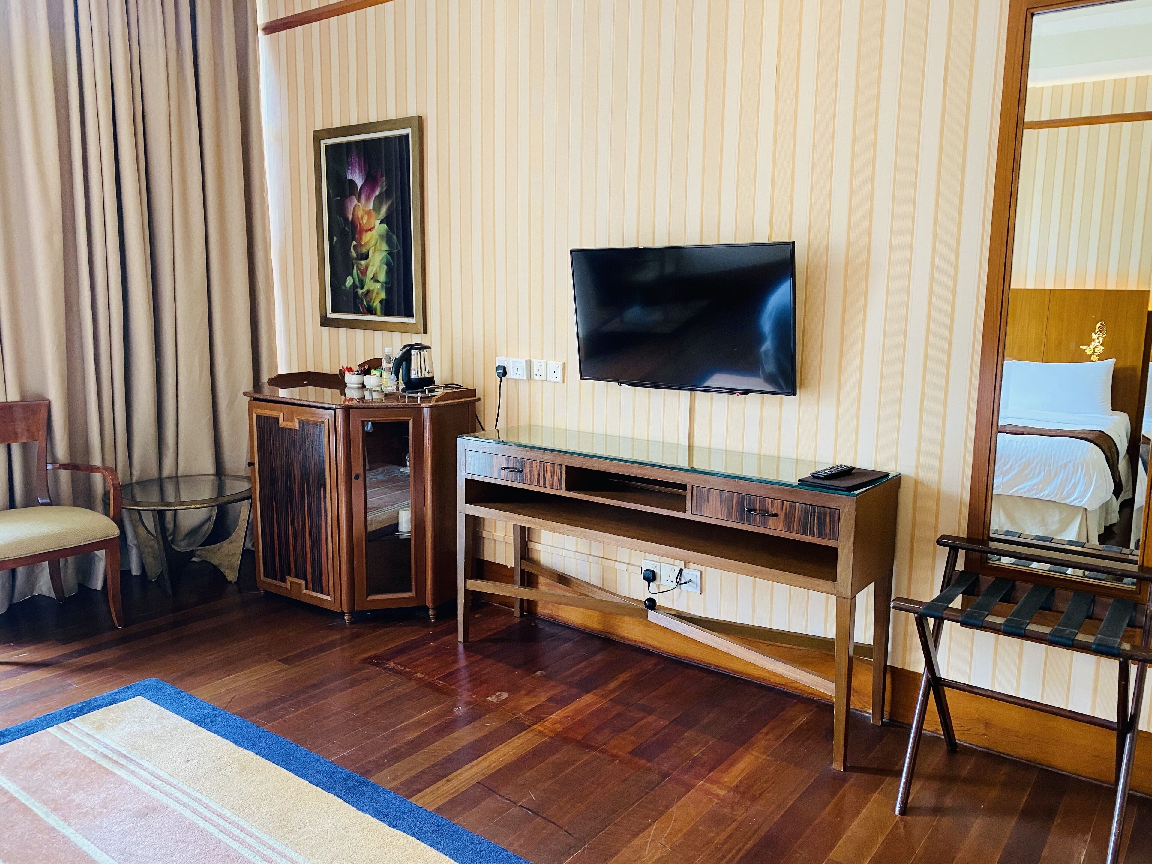 A hotel room with a wooden floor, a glass table, a TV mounted on the wall, a bed with a white and brown bedspread, and a wooden chair.
