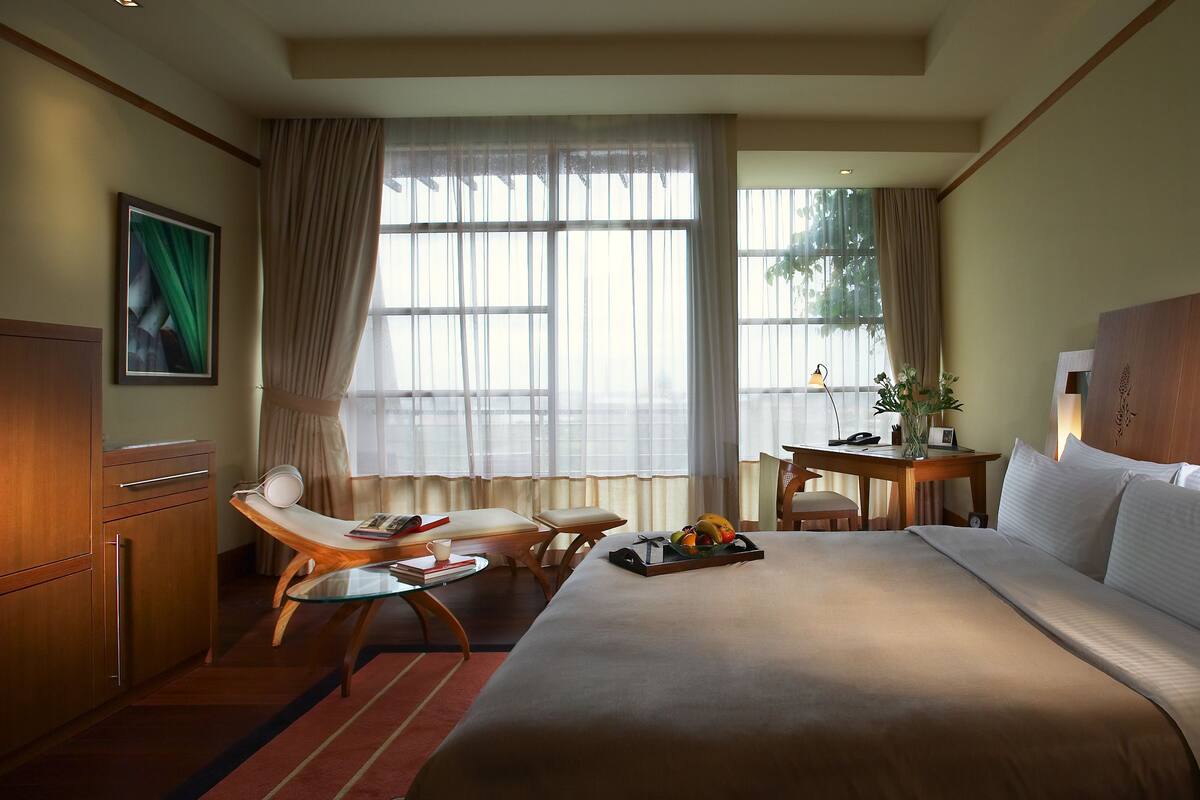 A hotel room with a large bed, a desk with a chair, a small table with a plant, and a window with curtains.