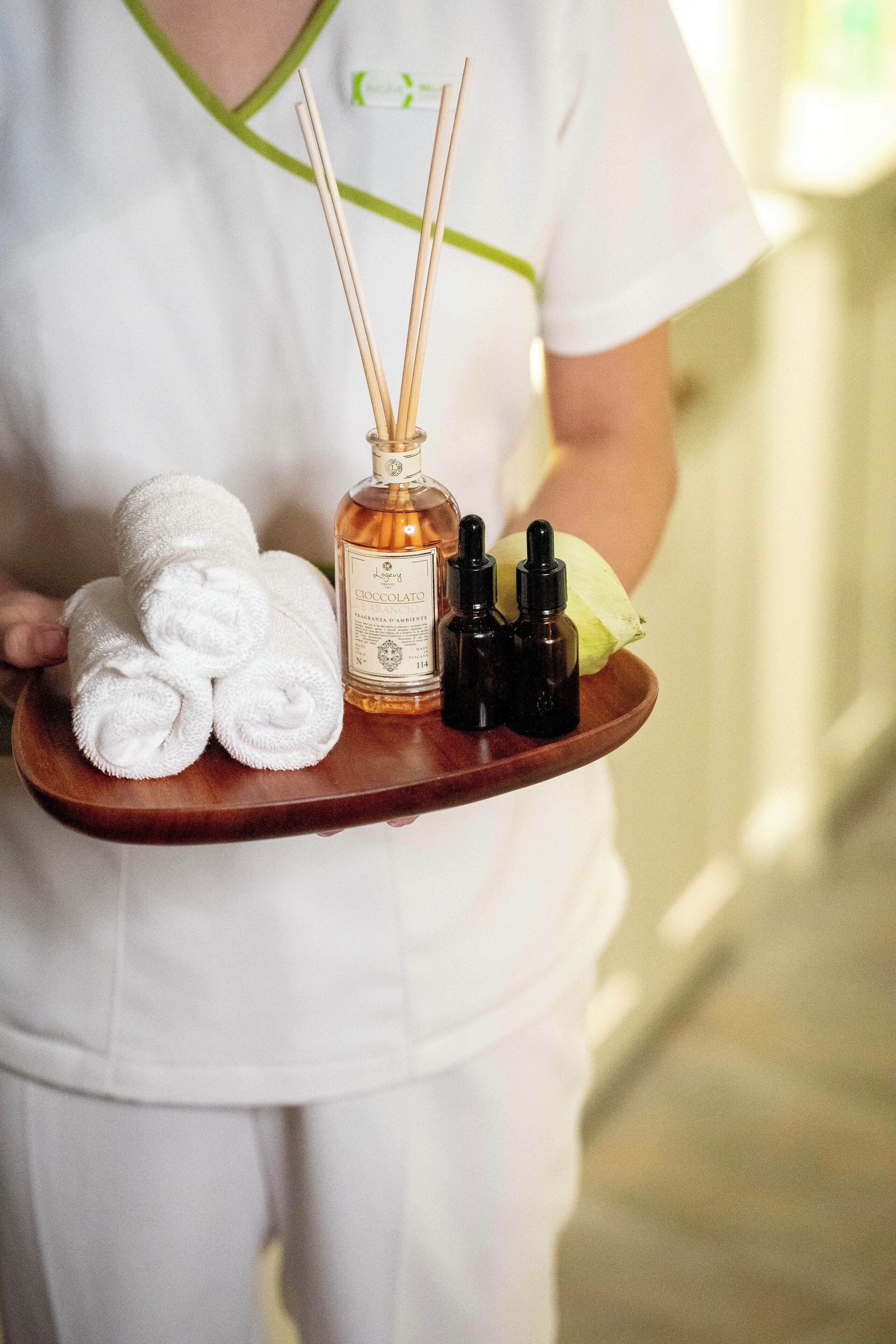A tray with rolled towels, a bottle of Caffè Latté, and two small bottles, likely containing essential oils, held by a person in a white uniform.