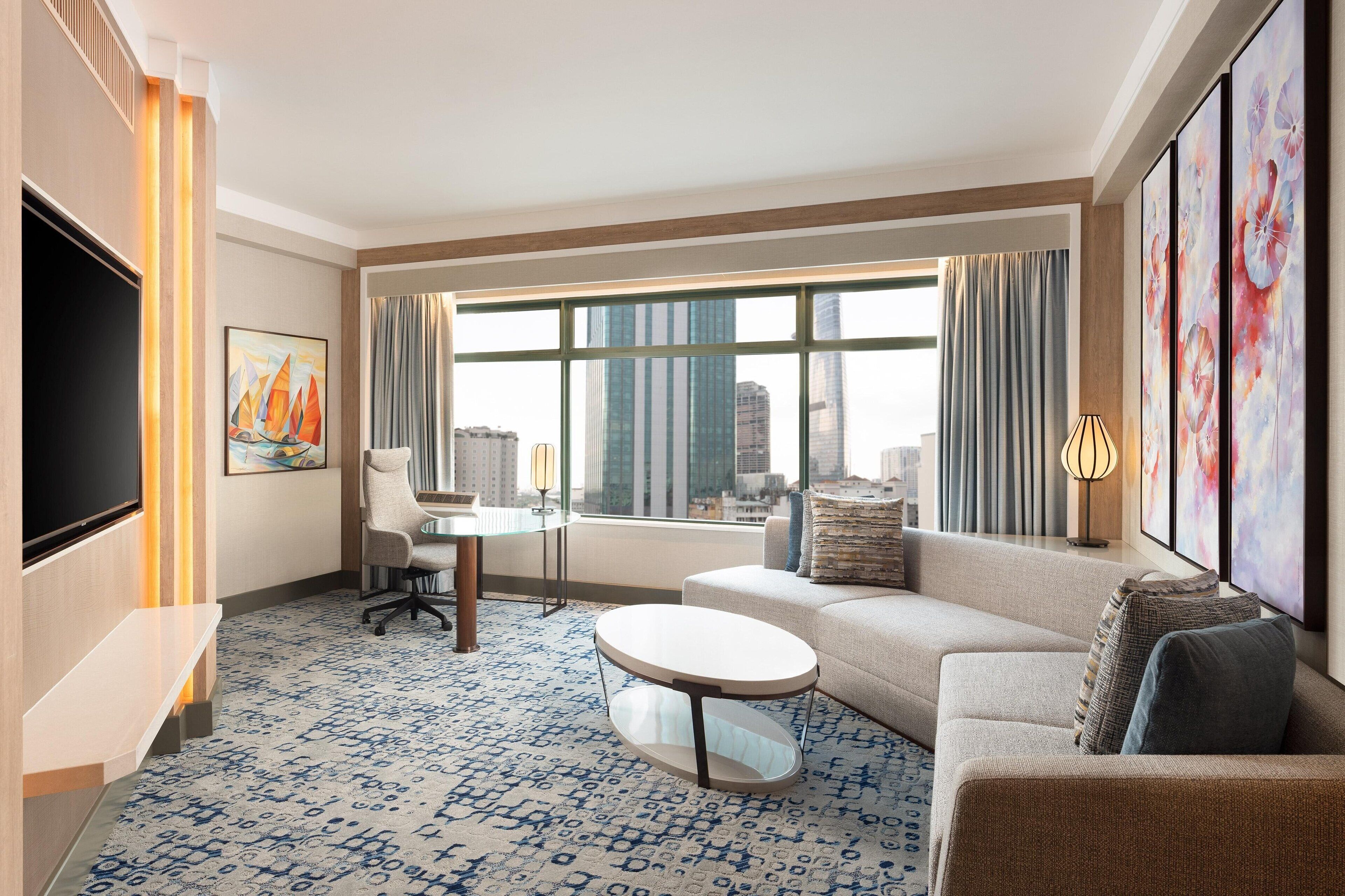 Une chambre d’hôtel moderne dotée d’une grande fenêtre, d’un téléviseur à écran plat, d’un canapé, d’une table basse et d’un bureau avec une chaise.