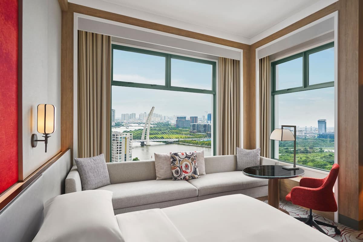 A hotel room with a large bed, a sofa, a small table, and a red chair. There are two large windows with a view of a cityscape and a bridge.