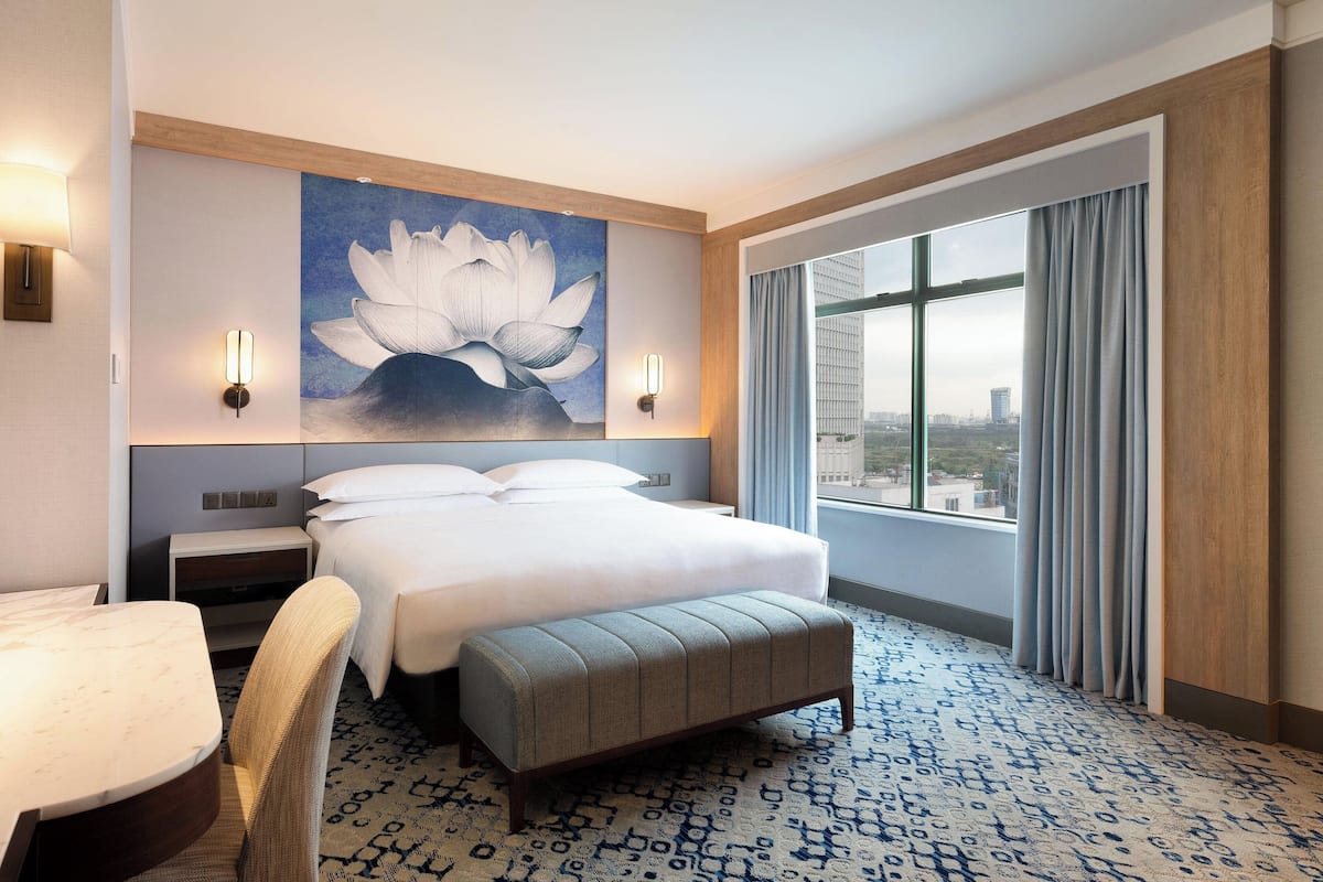 A hotel room with a large bed, a desk, a chair, a bench, a painting of a lotus flower, and a large window with curtains.