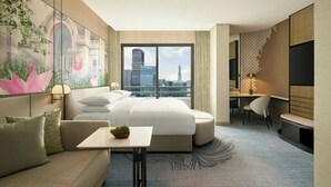 A modern hotel room with a large bed, a sofa, a small table with a plant, a desk, and a television. A large window offers a view of a cityscape.