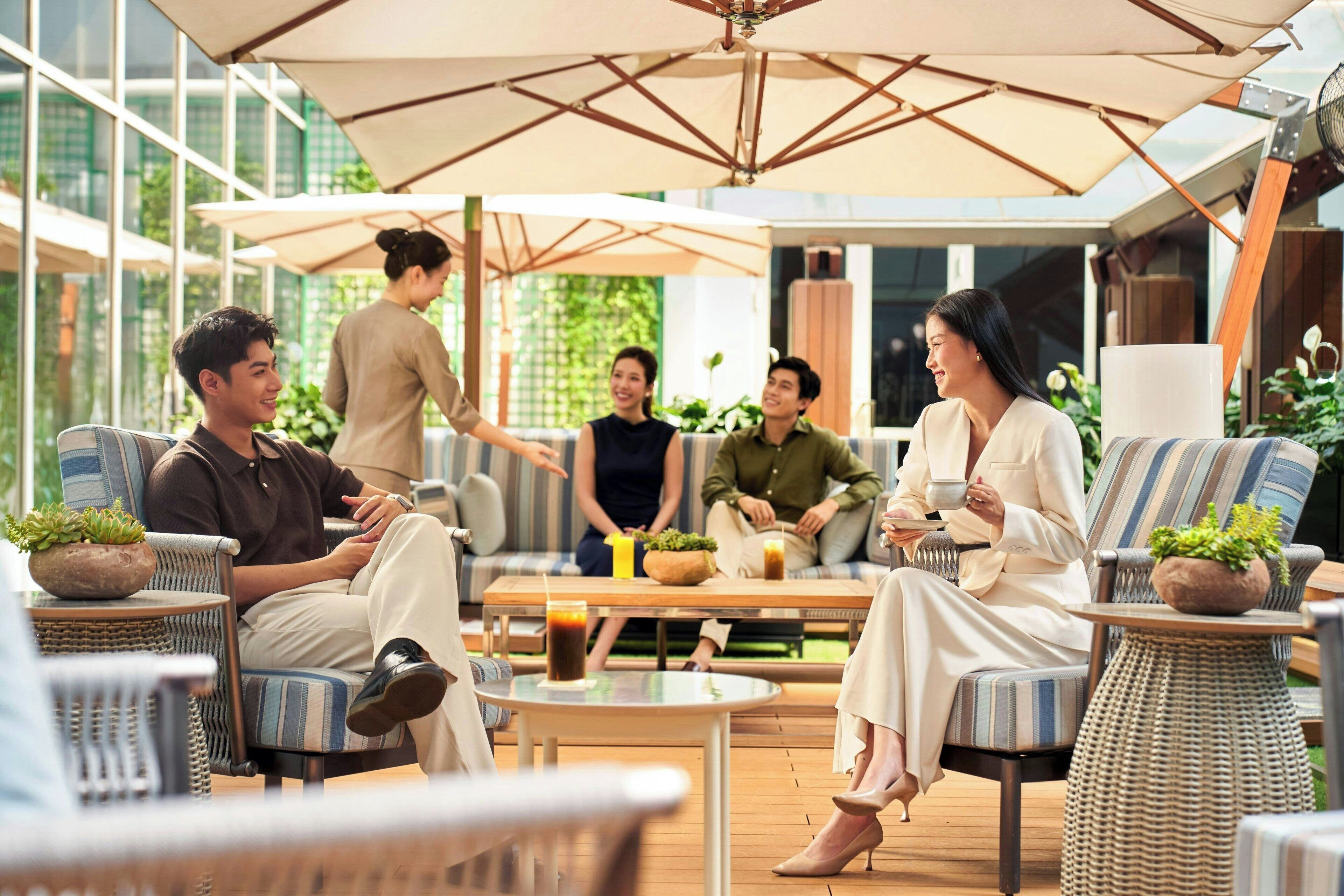 Un groupe de personnes est assis dans un espace de détente moderne en plein air, aménagé avec des meubles en osier, de grands parasols et des plantes en pot.