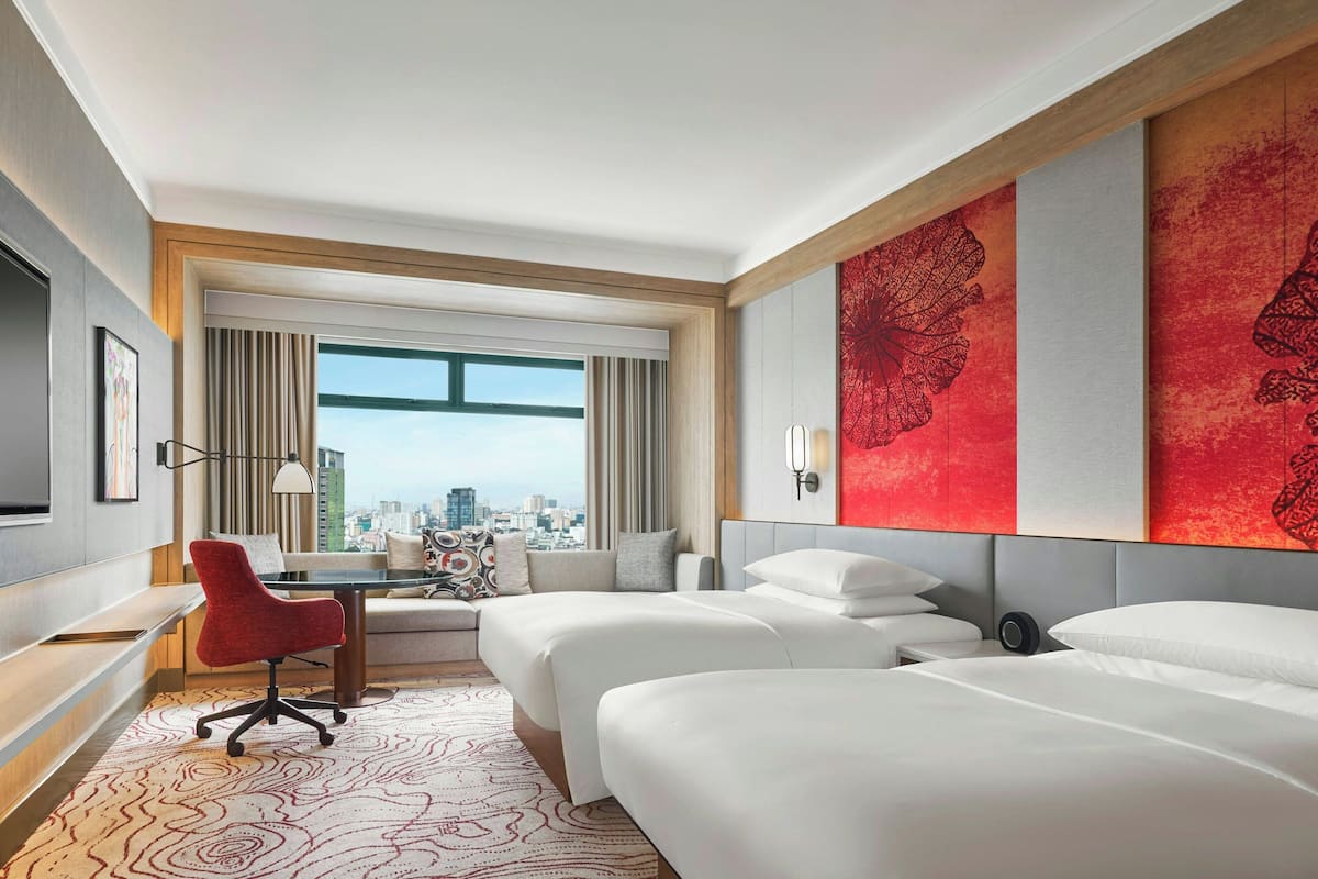 A hotel room with two beds, a desk, a red chair, and a large window offering a city view.