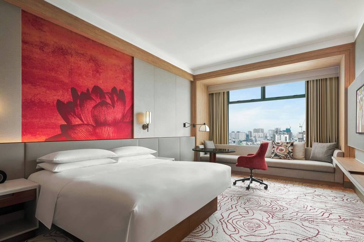 A hotel room with a large bed, a desk, a chair, a view of the city, and a red artwork on the wall.