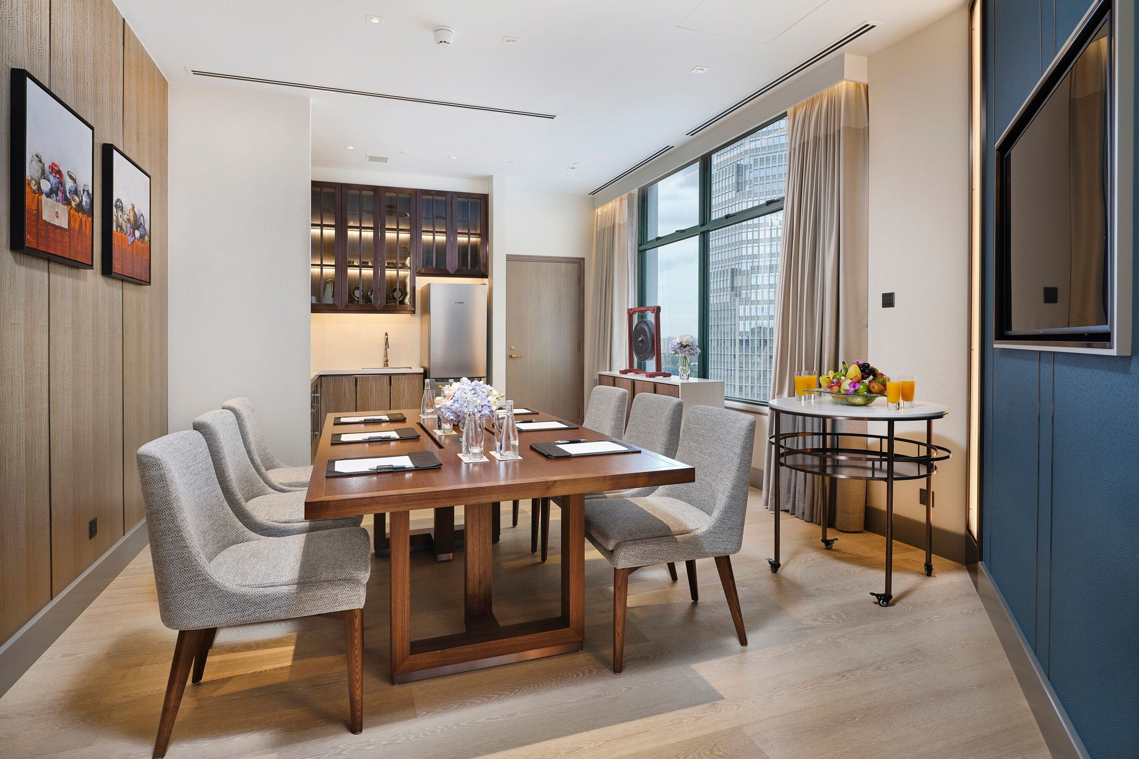 A modern dining area with a wooden table, grey upholstered chairs, and a small side table with a vase and fruit.