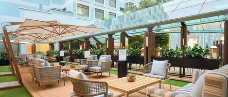 Un moderno espacio lounge al aire libre con mobiliario de mimbre, un dosel de vidrio y vistas a la ciudad.