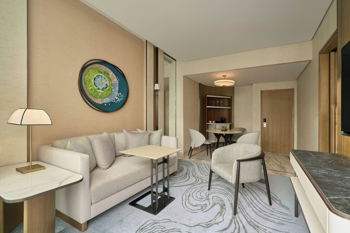 A modern hotel room with a sofa, a small table, a chair, a dining area, and a large painting on the wall.