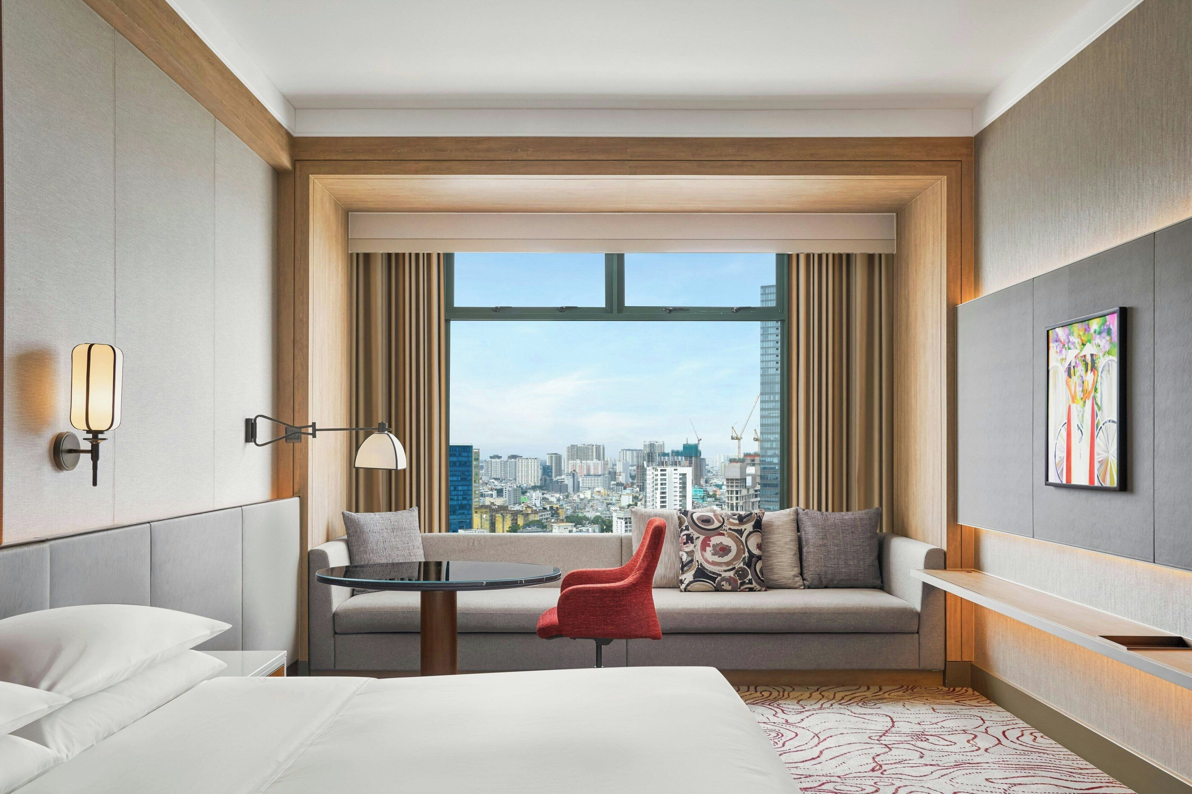 Habitación de hotel con cama, sofá, una silla roja, una mesita y un ventanal con vista a la ciudad.