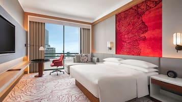 A hotel room with a large bed, a TV, a desk, and a chair. There is a large window with a view of the city.