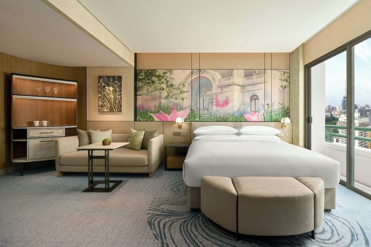 A modern hotel room with a large bed, a sofa, a small table, and a cabinet. There is a large window with a city view.