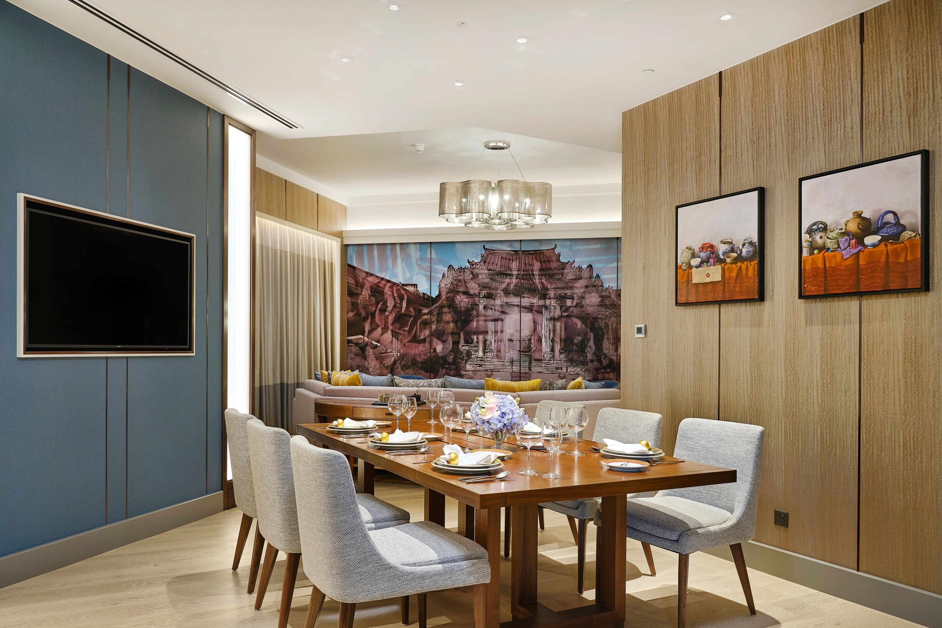 Une salle à manger moderne avec une table en bois dressée pour six personnes, entourée de chaises rembourrées. Un grand téléviseur à écran plat est fixé au mur, et deux œuvres d’art encadrées sont accrochées au-dessus de la table.