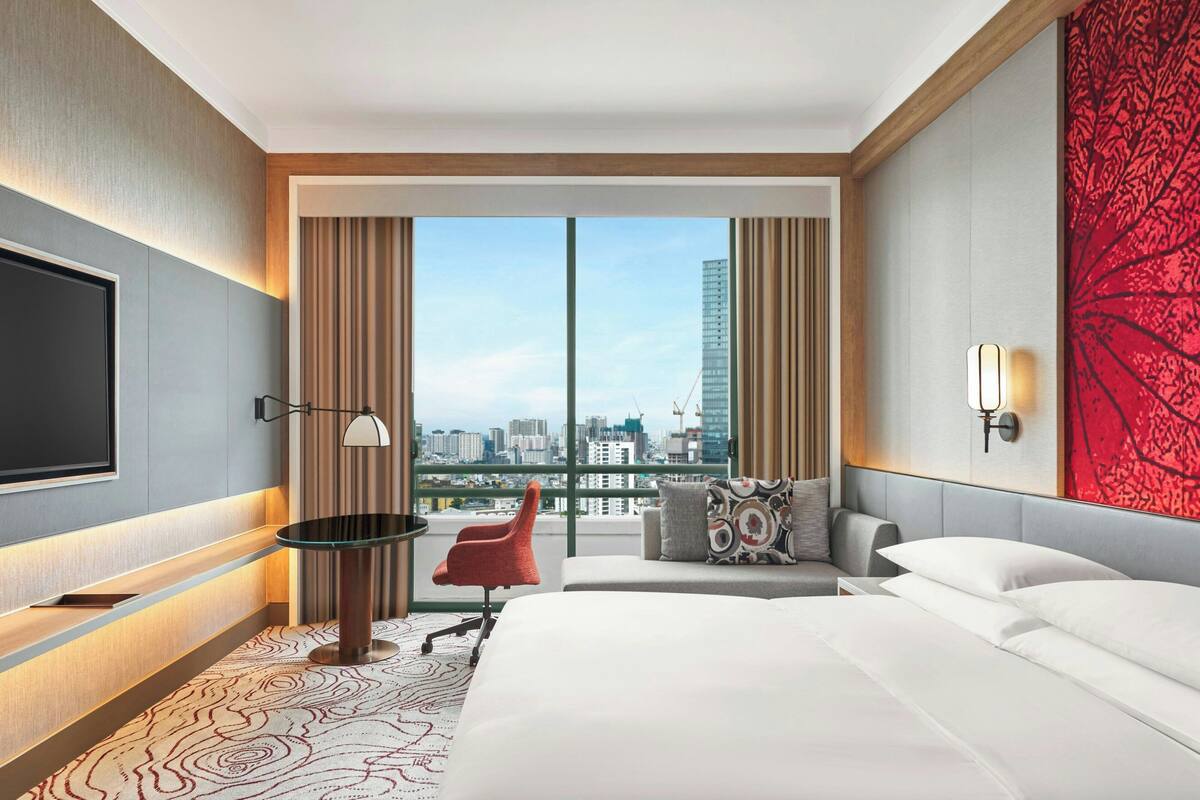 A modern hotel room with a large bed, a desk, a chair, and a TV. There is a large window with a city view.