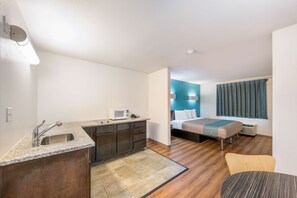 Una habitación de hotel compacta con kitchenette, una cama con cabecera azul, una mesita pequeña y una silla.