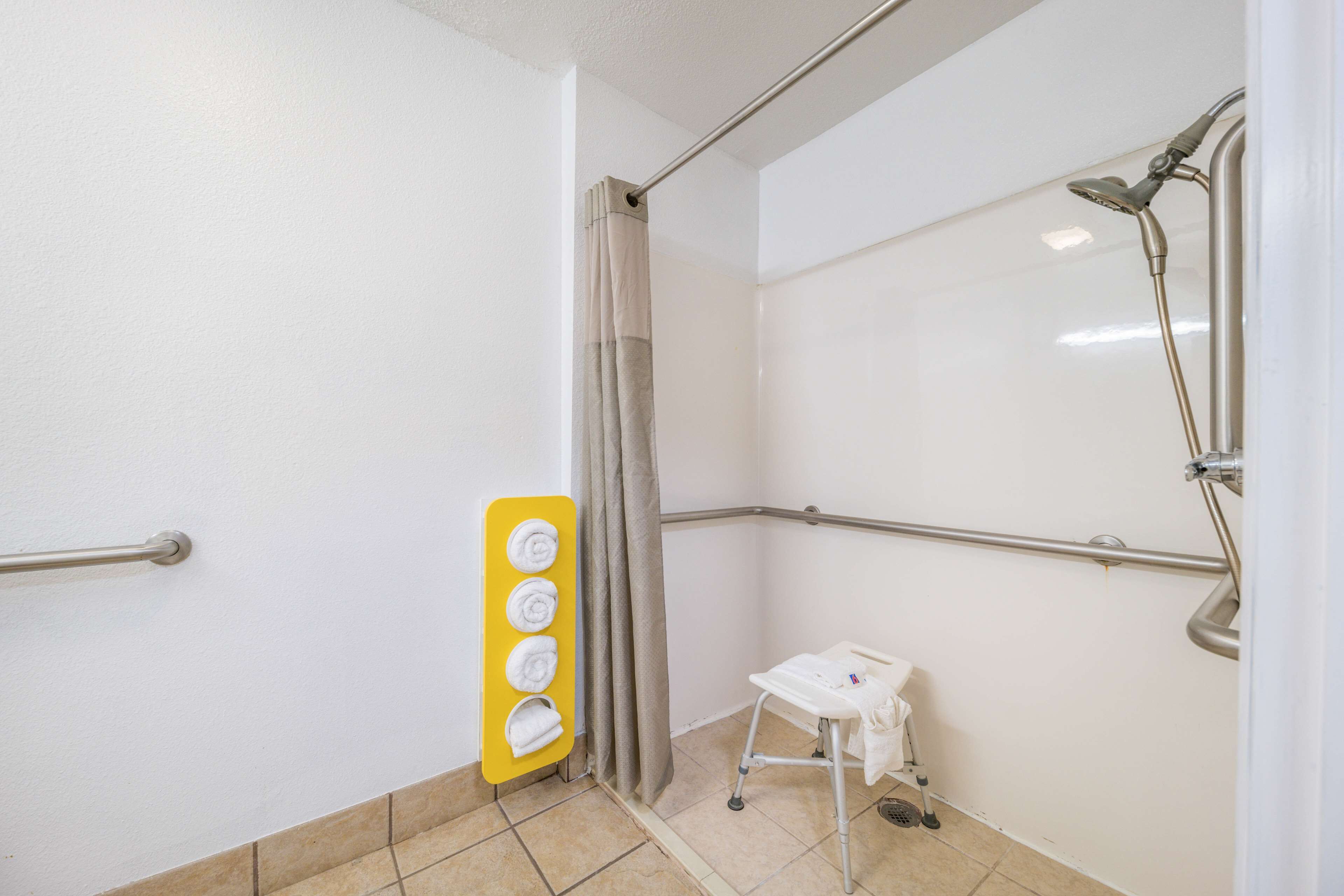 Uma área de chuveiro com barra de apoio, porta-toalhas amarelo e um banquinho branco pequeno.