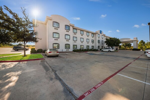 Un hotel con aparcamiento, varios coches y cielo despejado.