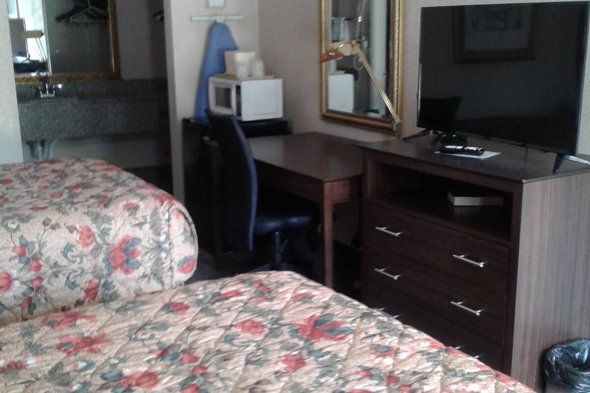 A hotel room with a bed, a desk, a chair, a television, a mirror, and a trash bin.