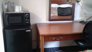 Ein kleines Hotelzimmer mit einem Holztisch, einem schwarzen Kühlschrank, einer Mikrowelle, einem einzelnen Sessel und einem Bett mit floralem Überwurf.