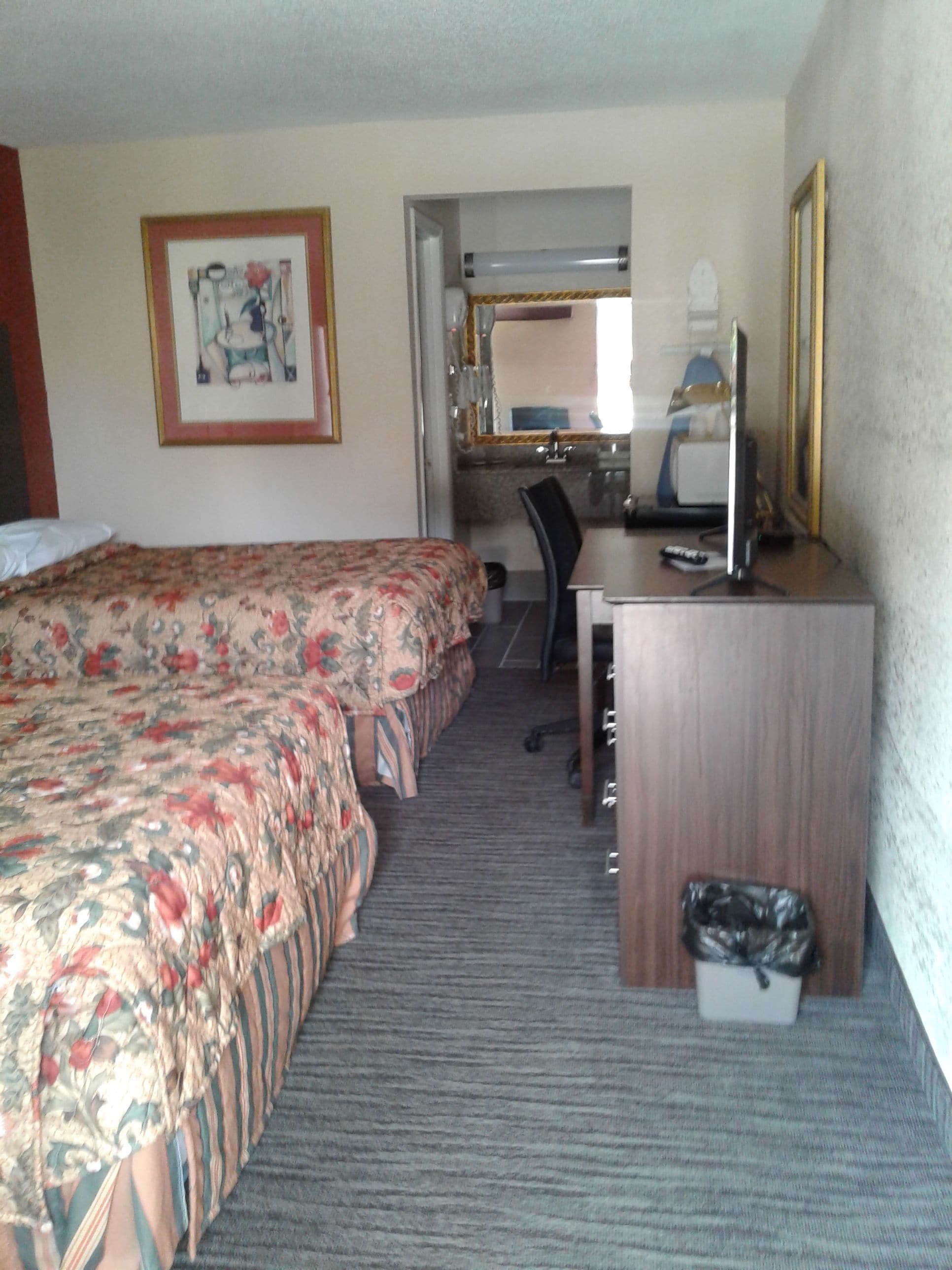 A hotel room with a bed, a desk, a chair, a computer, and a trash bin.