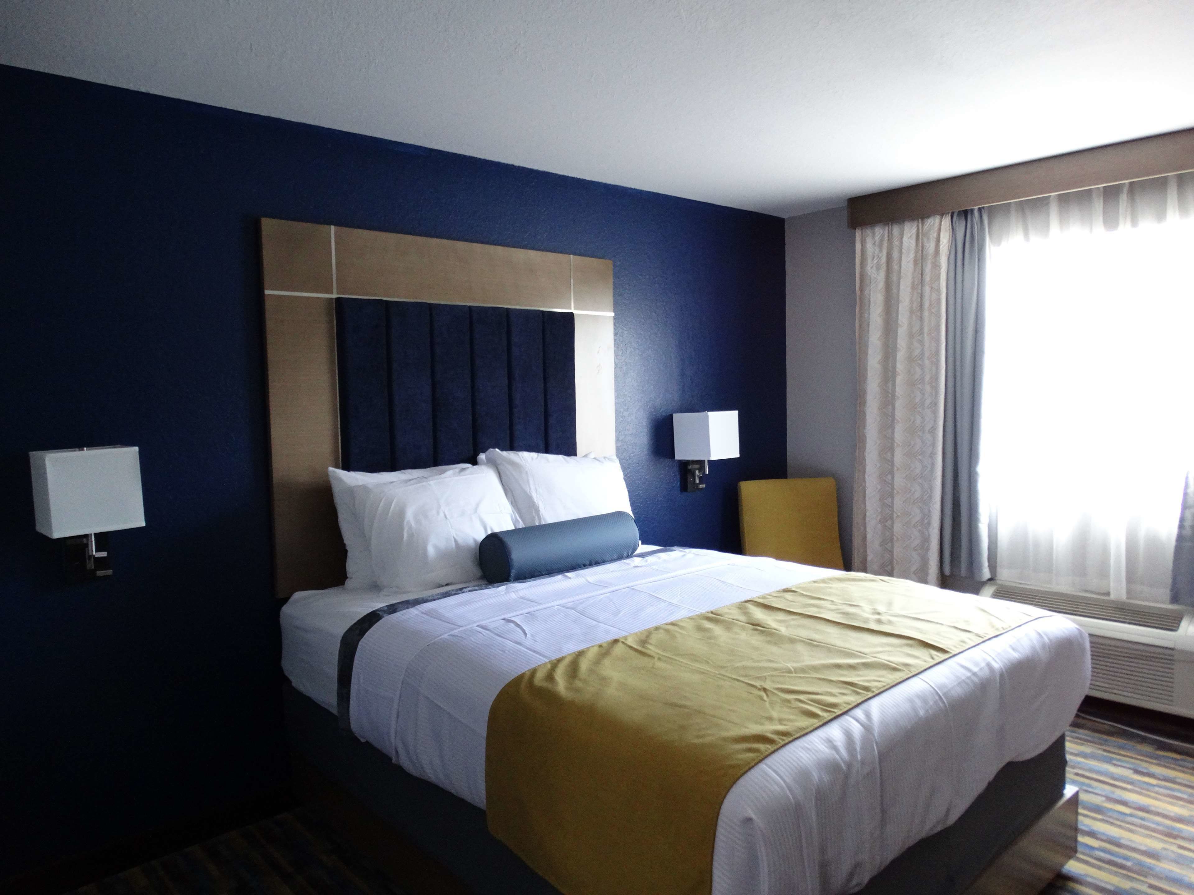 A hotel room with a large bed, two bedside tables with lamps, a window with curtains, and a wall-mounted air conditioning unit.