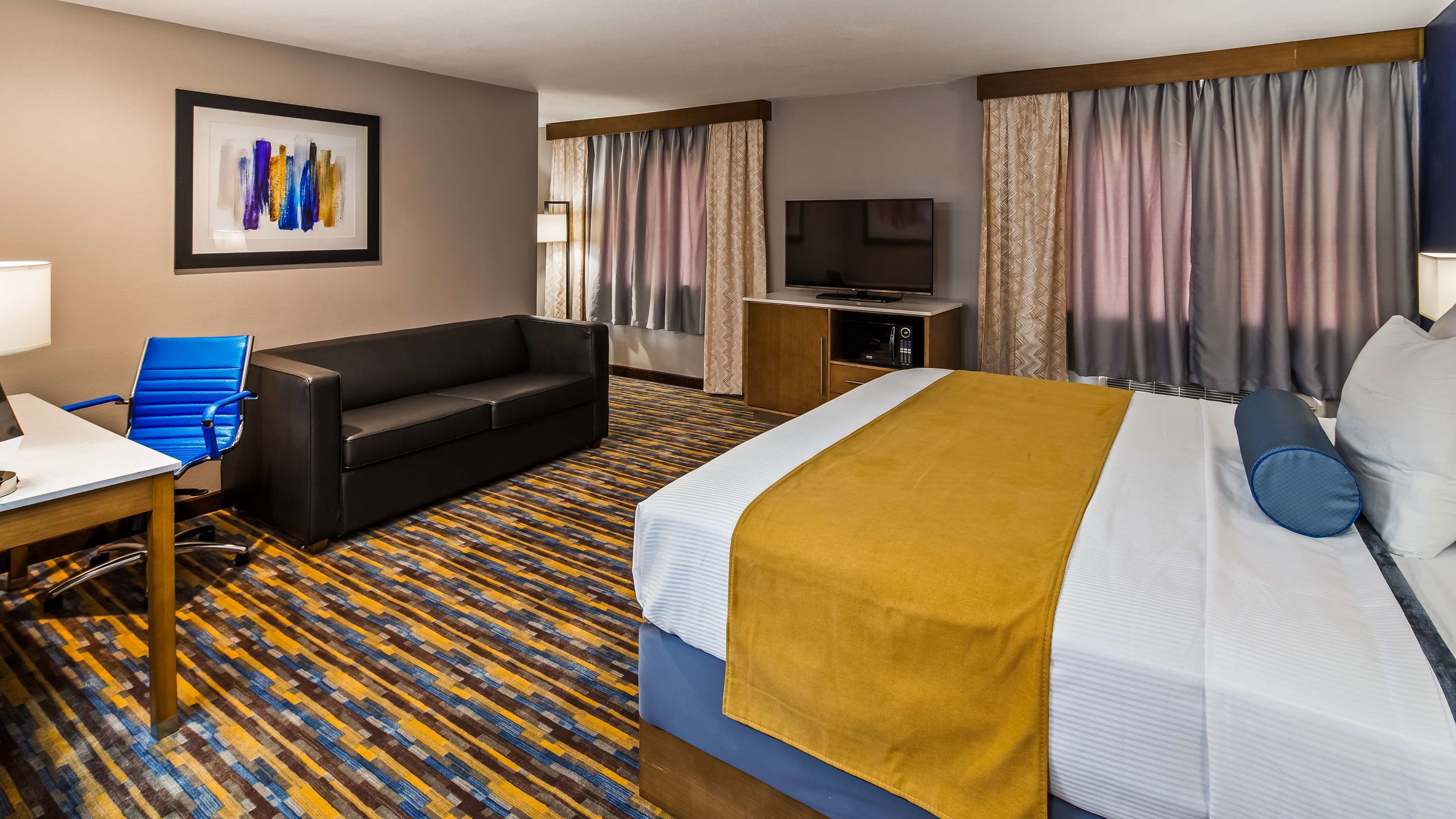 A hotel room with a bed, a sofa, a desk, a chair, a TV, and a painting on the wall.