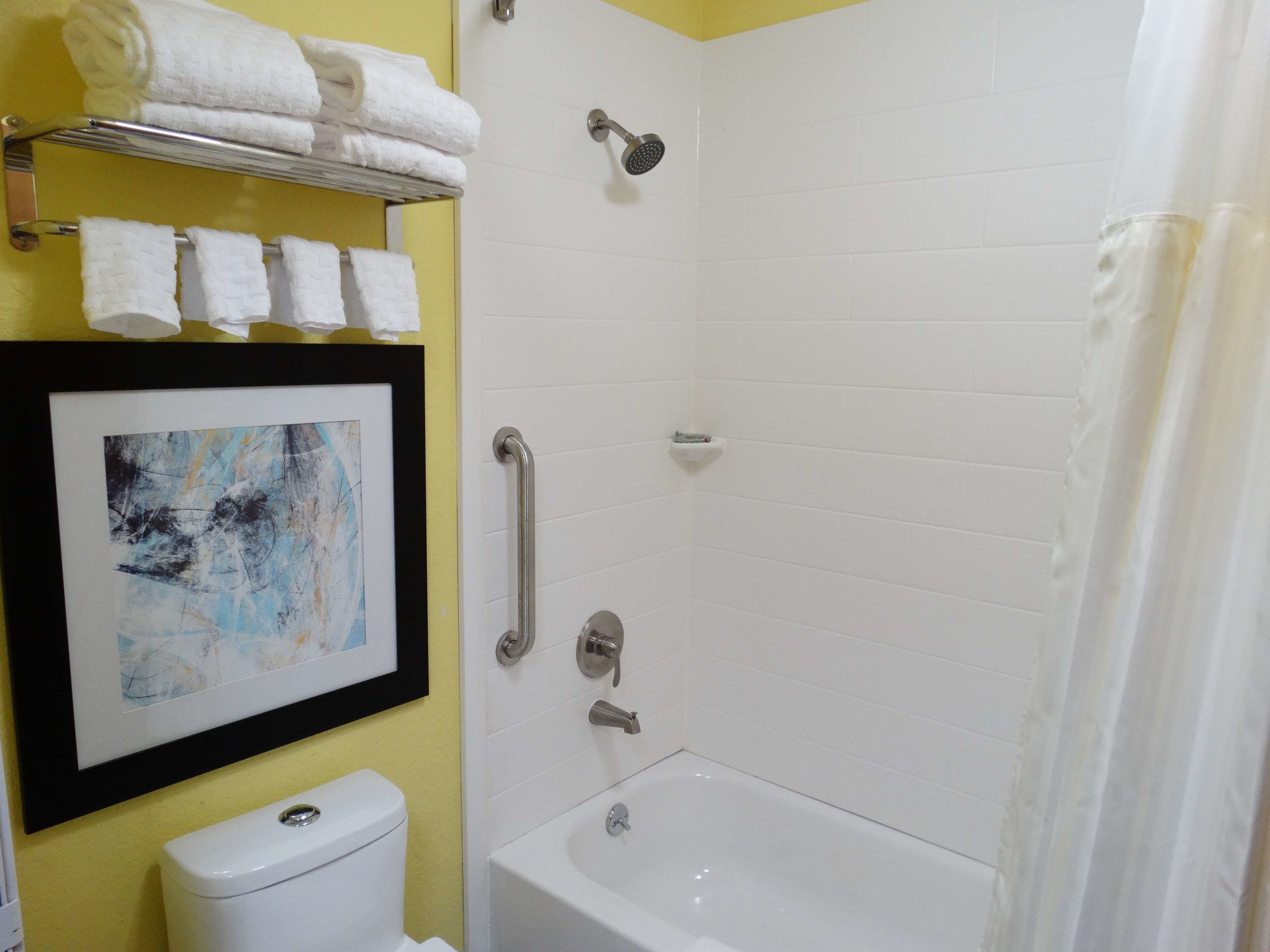 Un baño con una bañera blanca, un inodoro, un cuadro abstracto enmarco y un perchero con toallas dobladas.