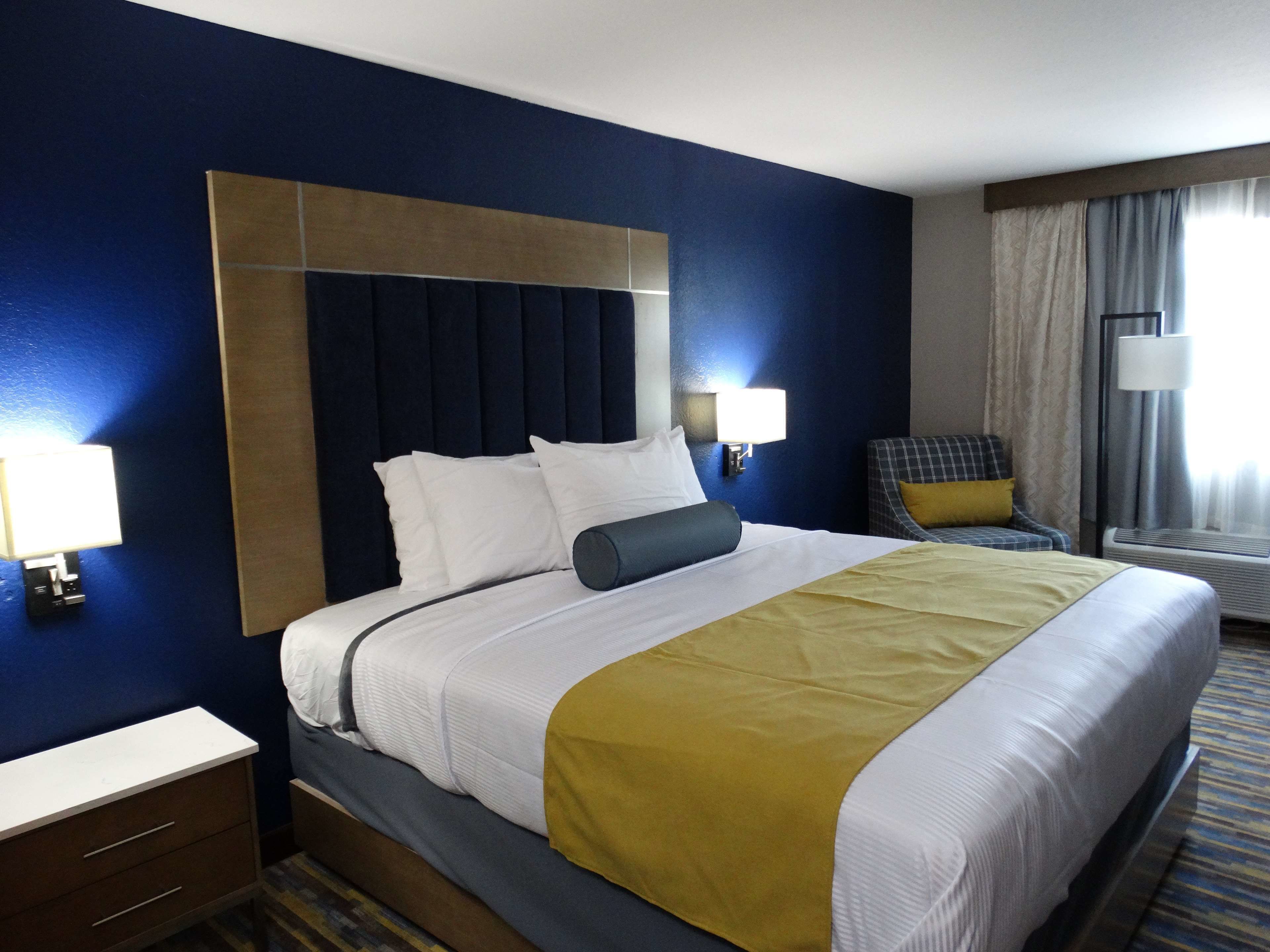 Habitación de hotel con una cama grande, una mesita de noche, una silla y un ventanal con cortinas.