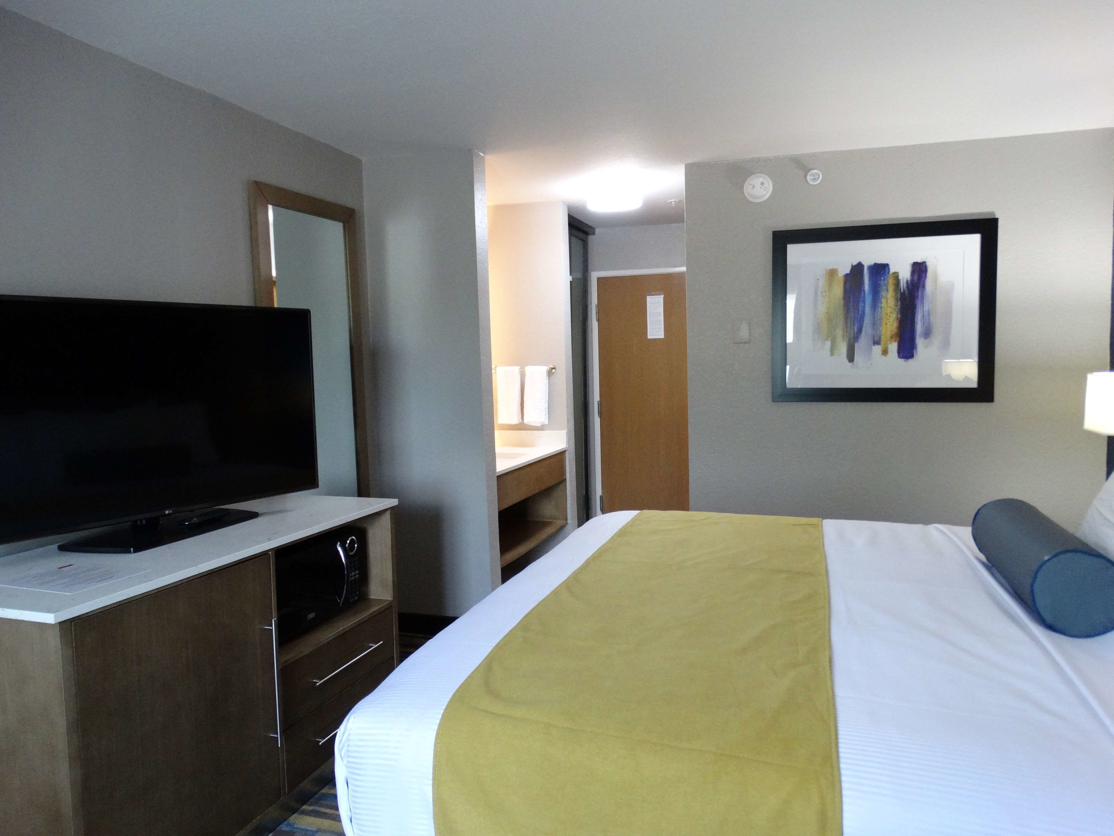 A hotel room with a bed, a TV on a stand, a desk, and a bathroom visible through an open door.