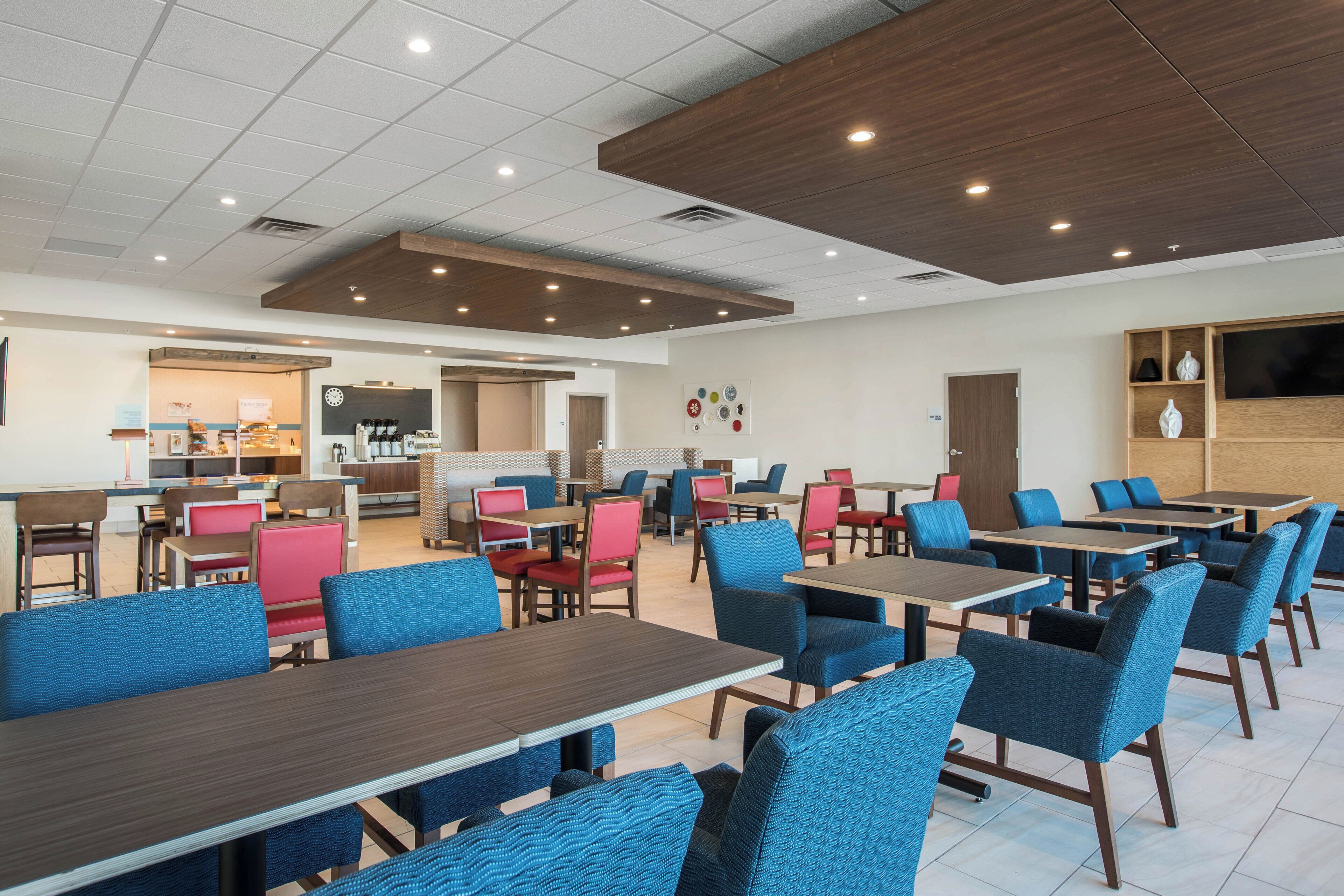 A modern dining area with multiple tables and chairs, a kitchen in the background, and a flat-screen TV mounted on the wall.