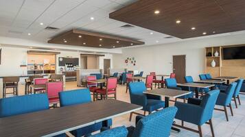 A modern dining area with multiple tables and chairs, a kitchen in the background, and a flat-screen TV mounted on the wall.