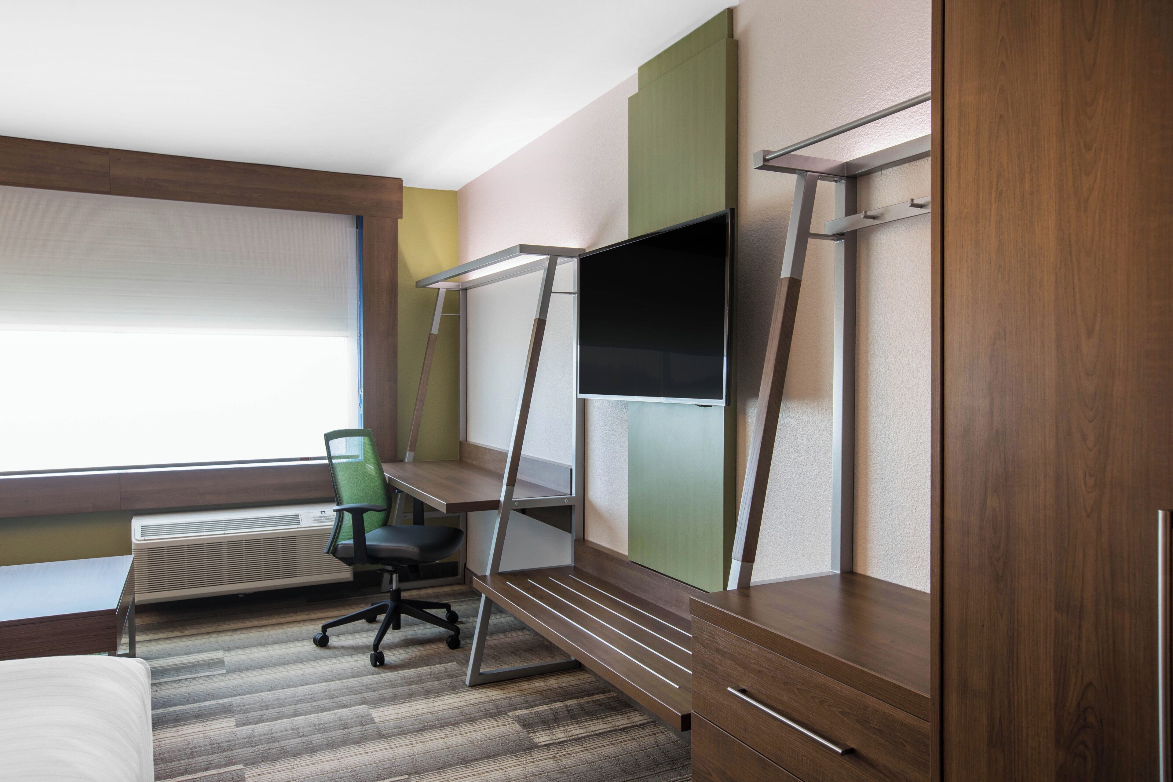 Habitación de hotel con cama, escritorio con silla, televisor montado en la pared y ventana con persianas.
