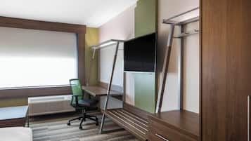A hotel room with a bed, a desk with a chair, a TV mounted on the wall, and a window with blinds.