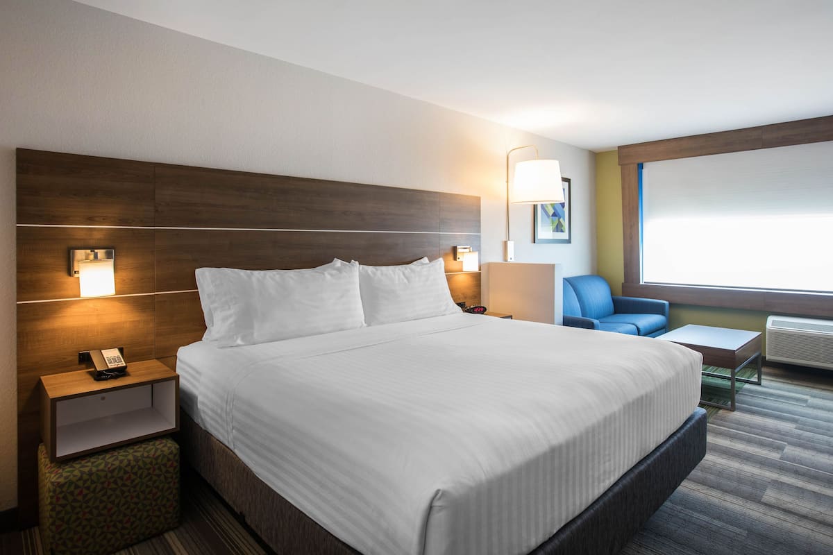 Una habitación de hotel con una cama grande, mesitas de noche, una silla, una mesita pequeña y una ventana con persianas.