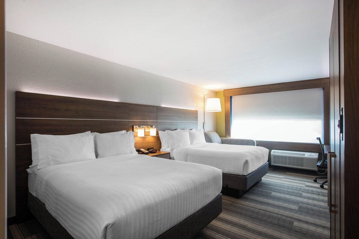 A hotel room with two beds, a headboard, bedside lamps, a window, and an air conditioning unit.