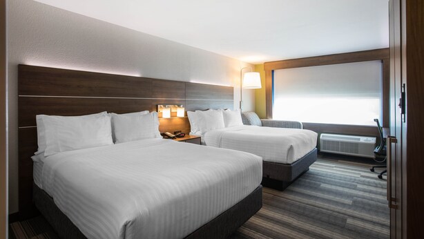 Habitación de hotel con dos camas, una mesita de noche con lámparas, una ventana con persianas y un aire acondicionado de pared.
