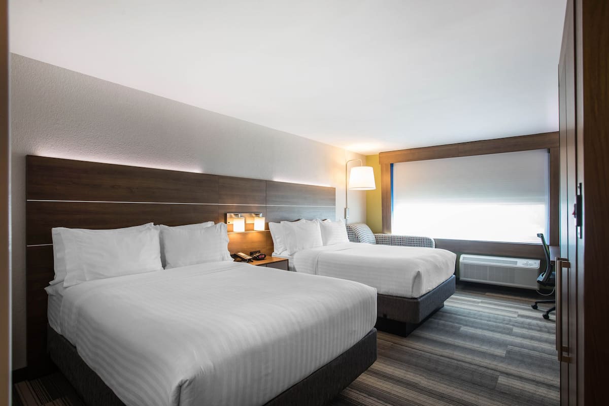 Habitación de hotel con dos camas, una mesita de noche con lámparas, una ventana con persianas y un aire acondicionado de pared.