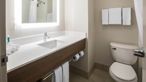 Un baño con lavamanos blanco, espejo y retrete. Hay toallas y artículos de aseo en el lavamanos.