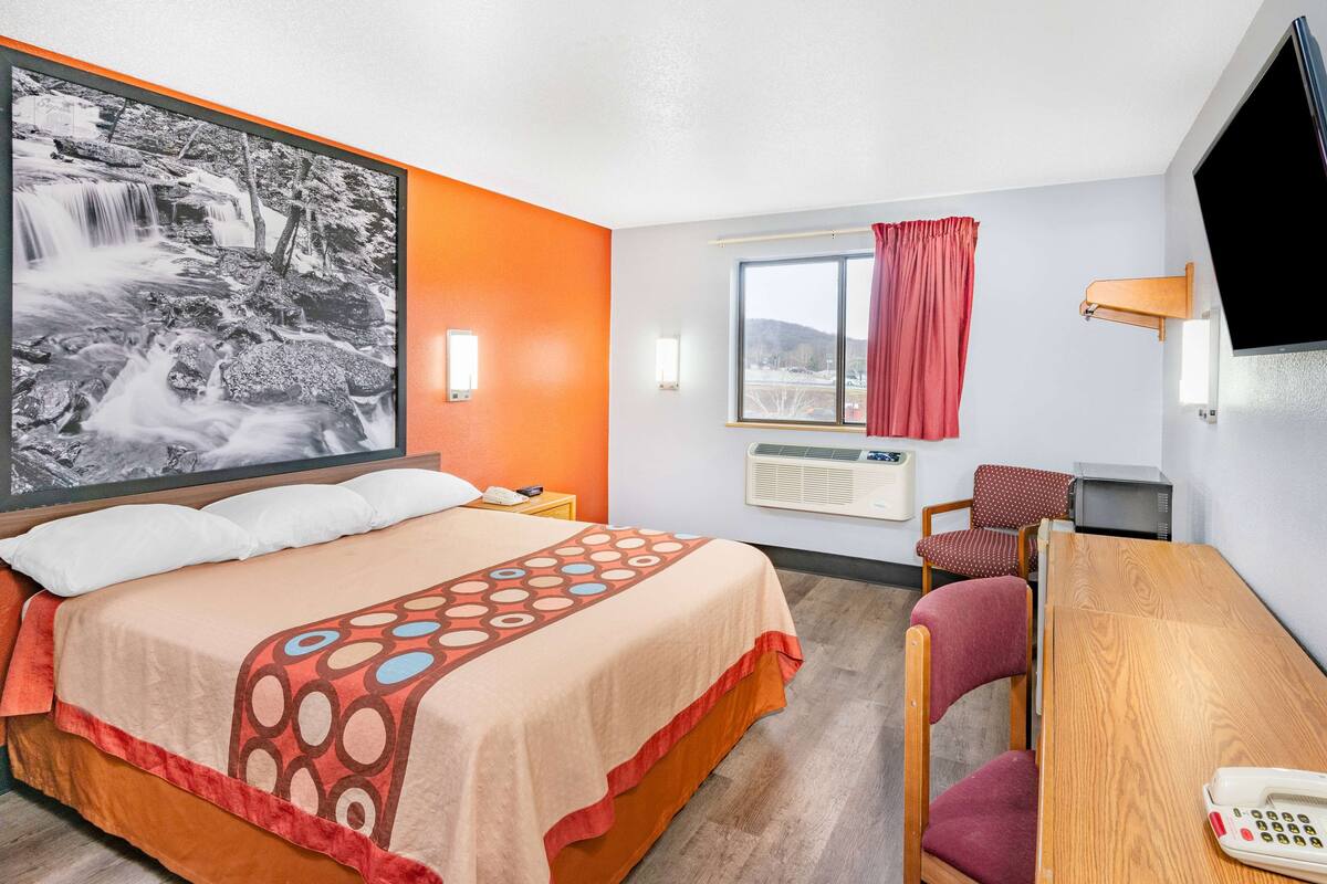 A hotel room with a bed, a desk, a chair, a TV, a window with curtains, and a wall-mounted air conditioner.