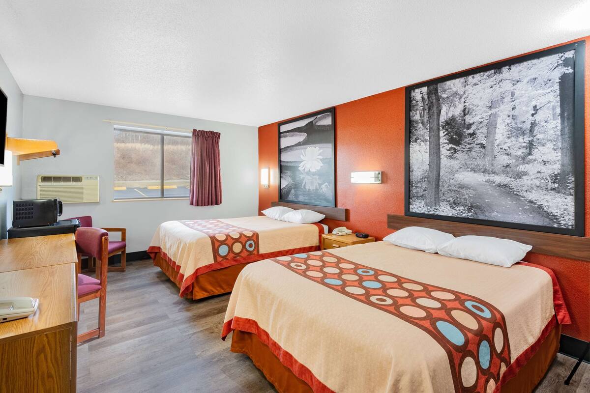 A hotel room with two beds, a desk, a chair, a window with curtains, a wall-mounted air conditioner, and a large framed picture of a forest path.