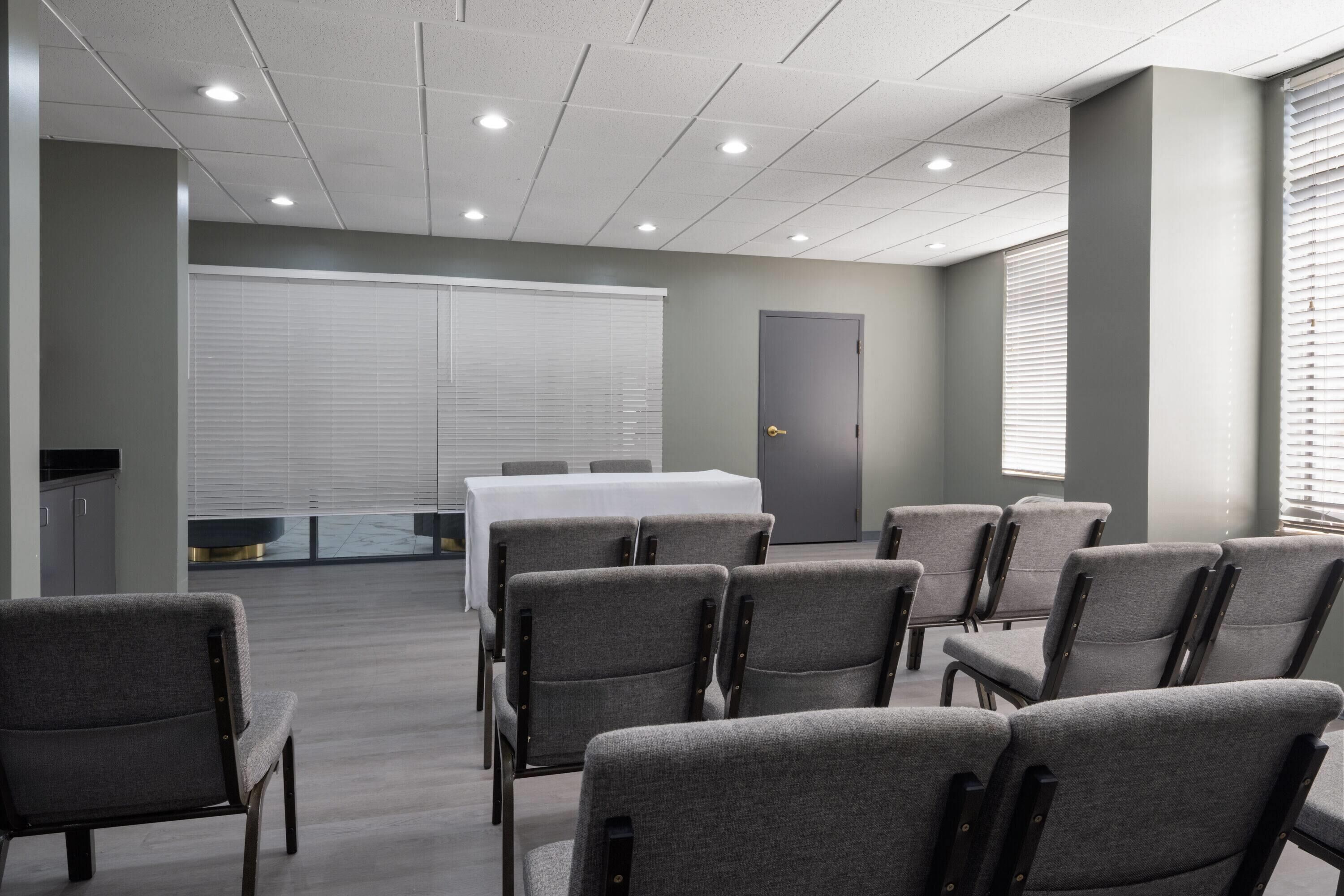 A conference room with grey chairs, a white table, and a large window with blinds.