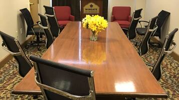 Un salón de conferencias con una larga mesa de madera, sillas de malla negra y sillas tapizadas de rojo. Se ve un logotipo en la pared y un jarrón con flores amarillas.