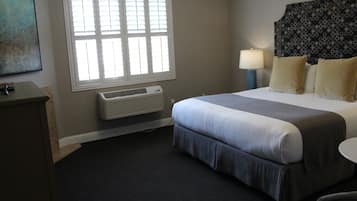 A hotel room with a bed, bedside lamp, air conditioning unit, and a window with blinds.