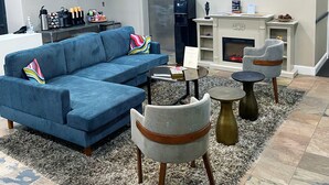 En moderne stue med en blå hjørnesofa, en grå lænestol og et rundt sofabord. Der er en pejs med en pejsestav og et indrammet billede over den.
