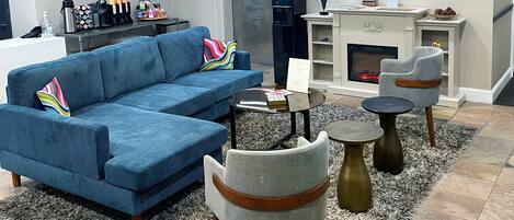 A modern living room with a blue sectional sofa, a grey armchair, and a round coffee table. There is a fireplace with a mantel and a framed picture above it.