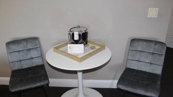 Uma pequena mesa redonda com base branca, acompanhada por duas cadeiras cinzas revestidas. Um jarro de vidro e um cartão estão sobre a mesa.