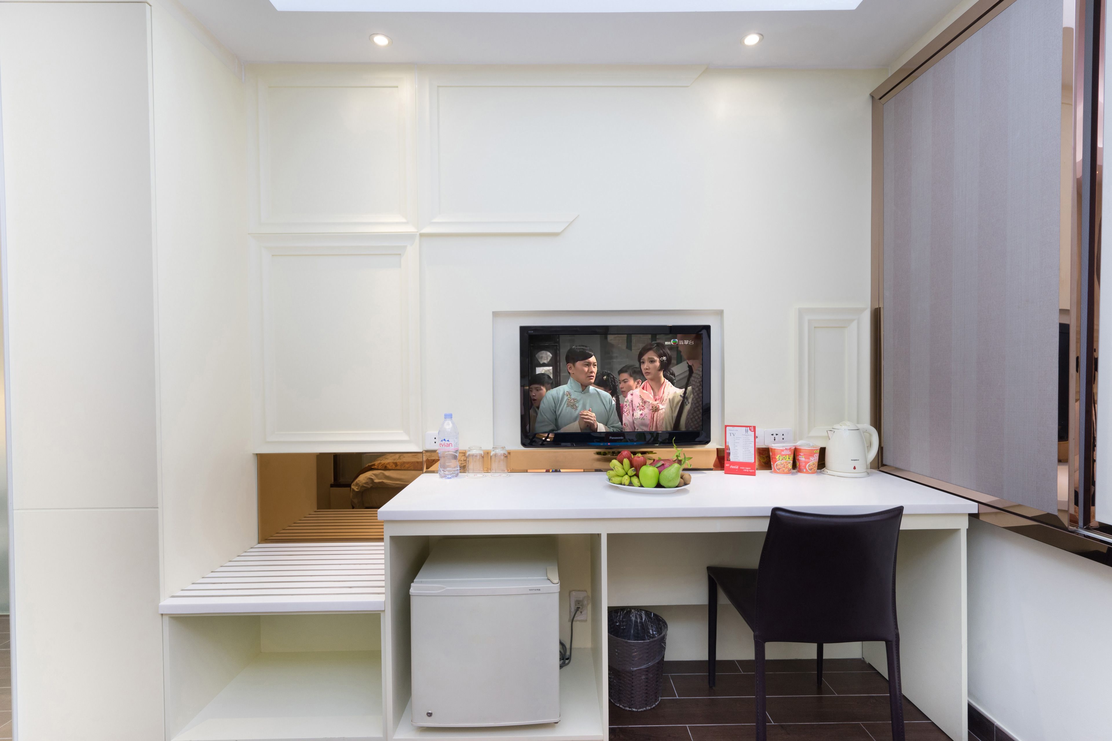 A modern room with a white desk, a television, a small refrigerator, and a black chair.